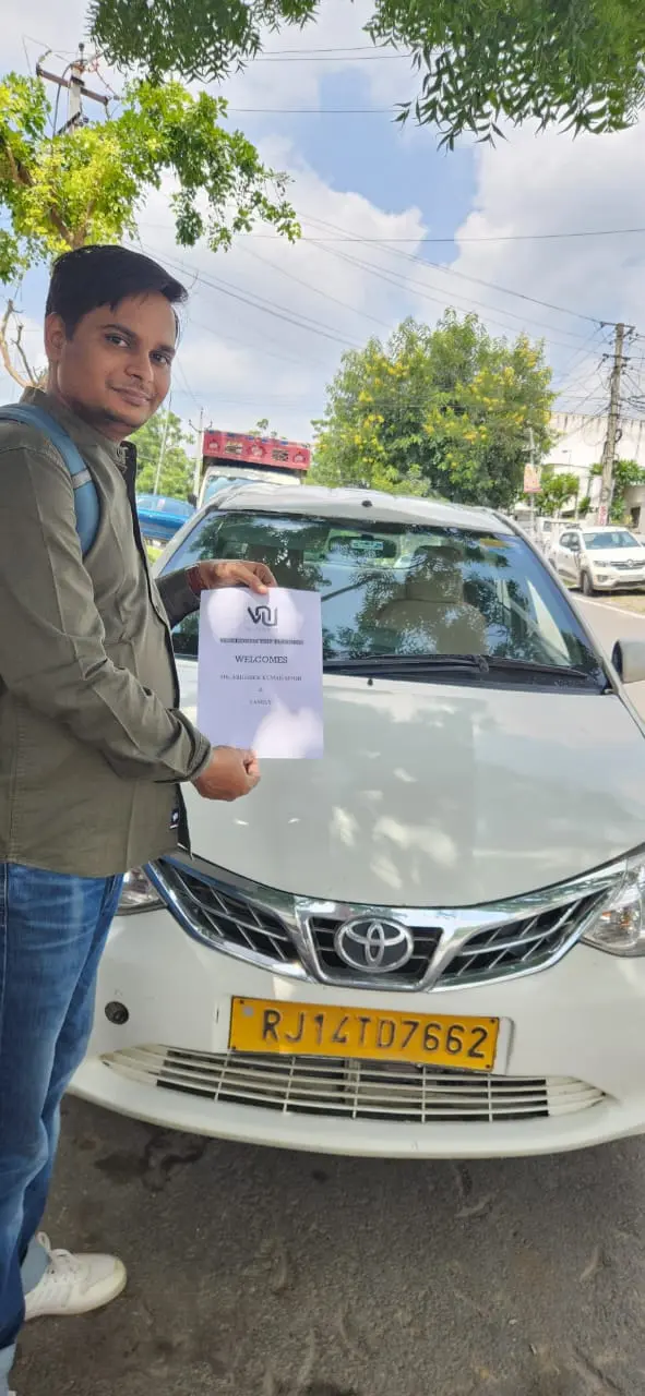 A man in a green shirt and blue jeans stands next to a white Toyota Etios with a yellow Rajasthan license plate, holding a PLA card of Vacationum..