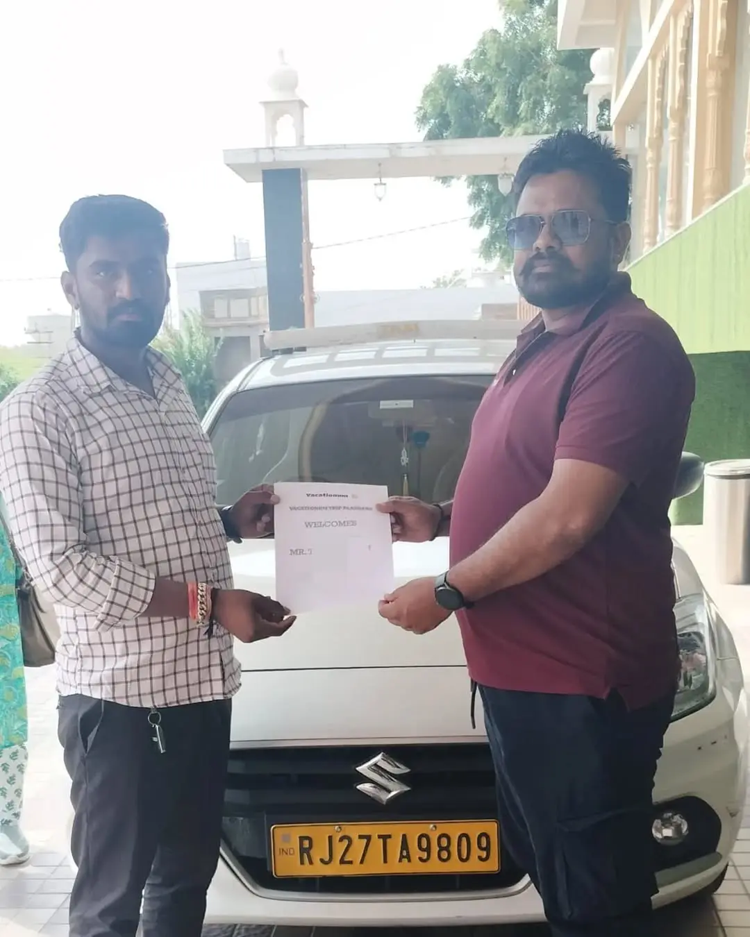 Guest standing in front of a white Suzuki car, holding a Vacationum's PLA card.