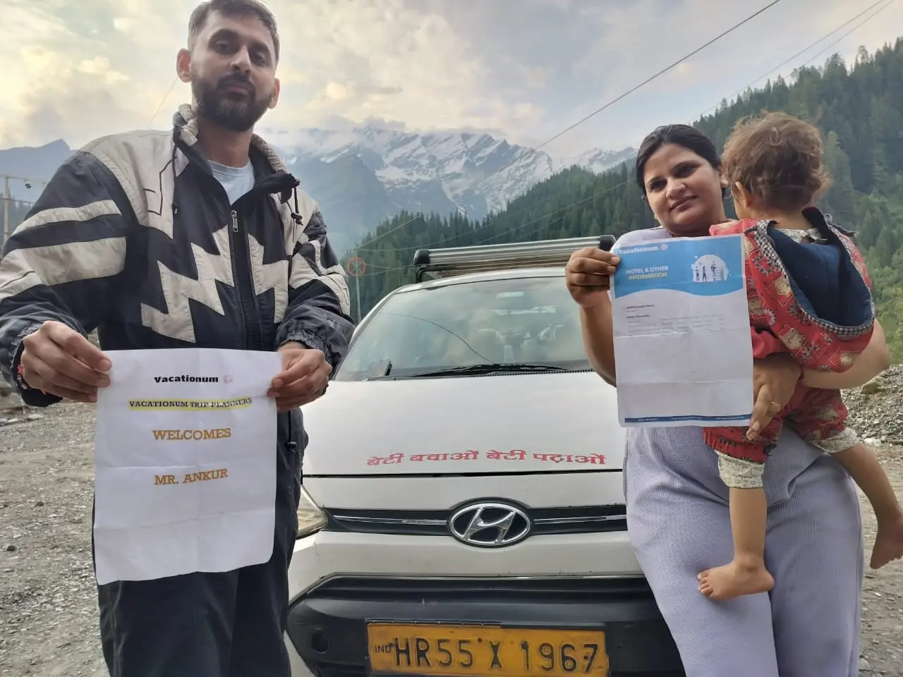 A man and a woman with a child pose in front of a white Hyundai hatchback in a snow-capped mountain region of India, holding Vacationum well planned itinerary and a welcome card.