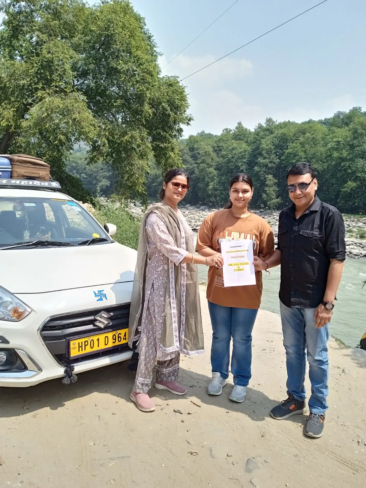 A family of three stands by a white Suzuki car on a riverbank in Shimla, Himachal Pradesh.