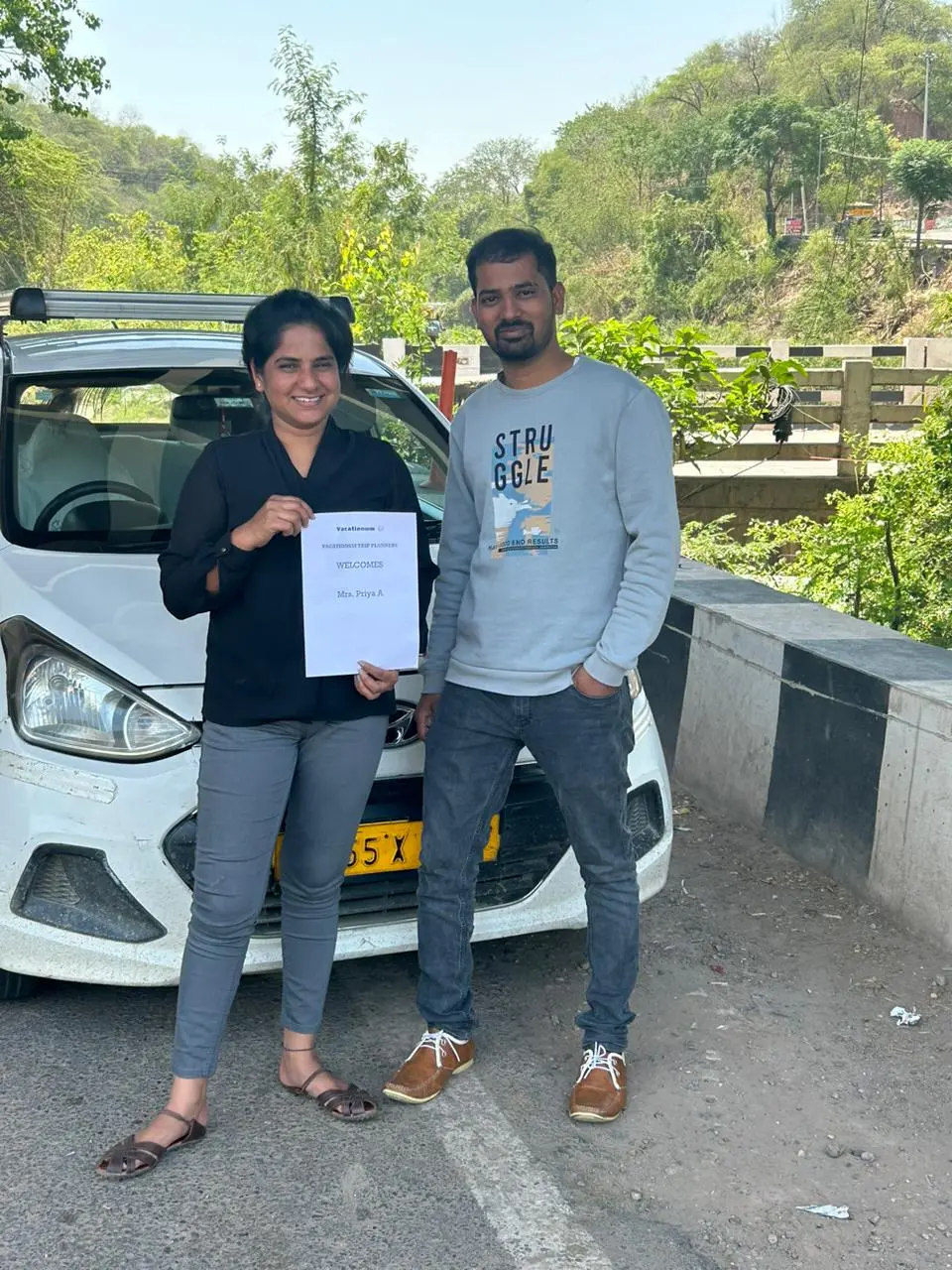 A couple stand together in front of a white car on a paved road, with the woman holding a welcome sign of Vacationum. Trees and a hillside are visible in the background.