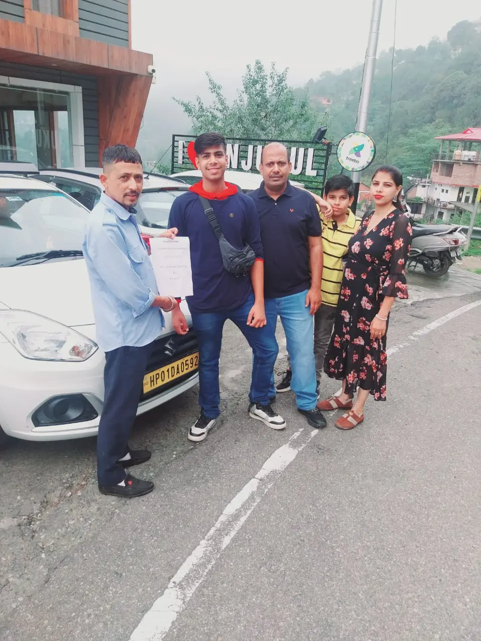A group of five people posing for a photo next to a white car on a mountain road in Khajjiar, Himachal Pradesh, with a lush green hillside and local signs in the background.