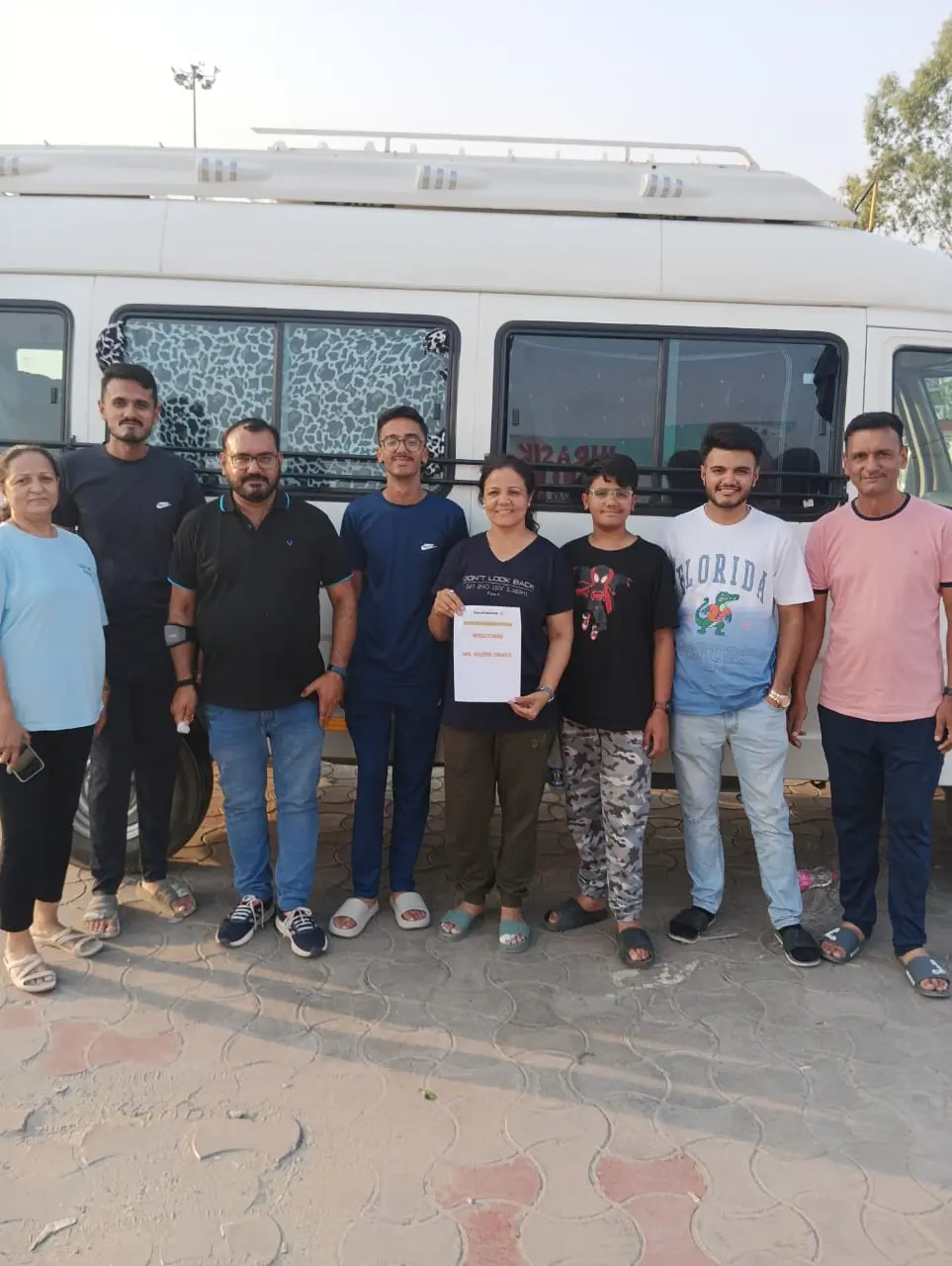 A group of nine people stands together for a group photo in front of a white tour bus, with one woman in the center holding a PLA card of Vacationum Trip Planners.