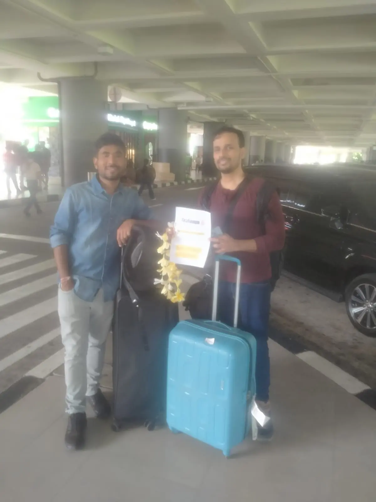 Two men posing with luggage and a floral lei at an airport terminal, with a "Malgudi" restaurant in the background at Bali Airport.