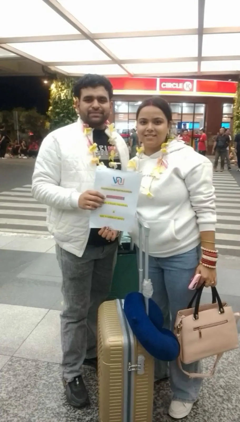 A smiling couple wearing flower garlands stands at an airport arrival area, holding a travel welcome sign near their luggage.