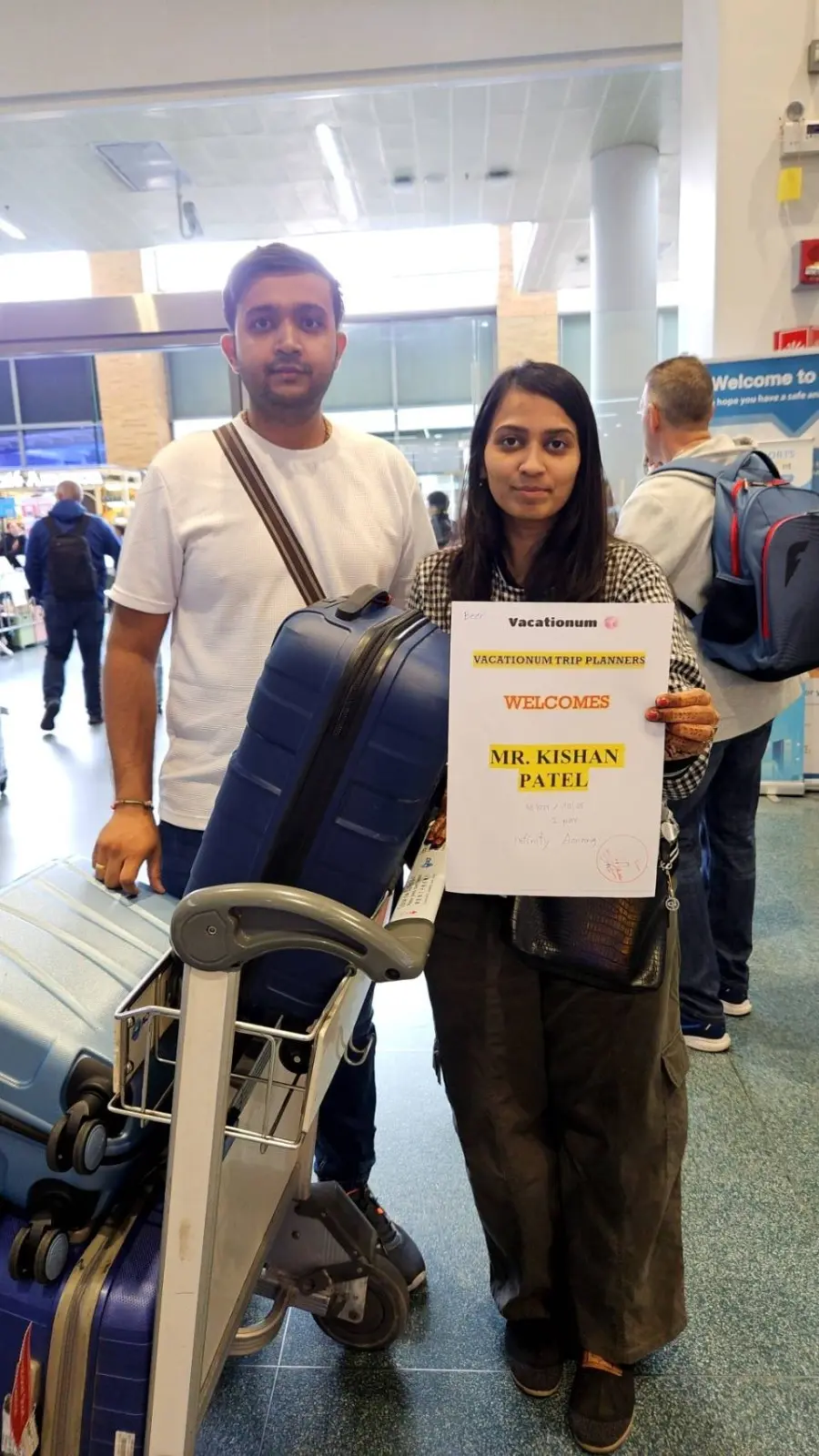 A man and woman standing in an airport; the woman holds a "Vacationum Trip Planners" sign welcoming next to a luggage cart with blue suitcases.