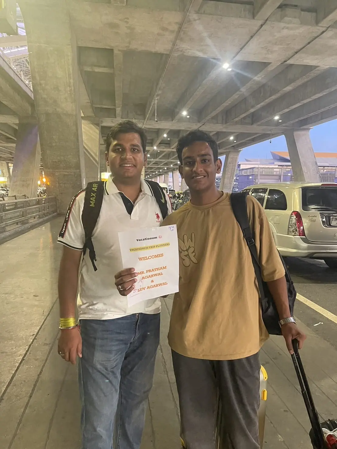 Two young boys at an airport arrivals area with a personalized welcome sign and luggage.