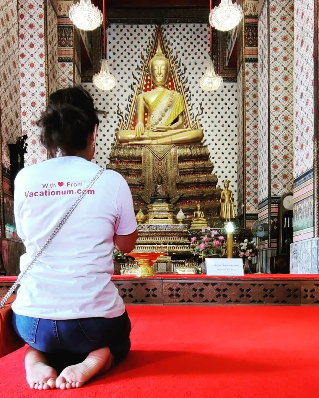 A woman kneels in prayer on a red carpet before a large, golden seated Buddha statue inside the ornate, patterned hall of Wat Arun in Bangkok, wearing cool Vacationum T-Shirt..